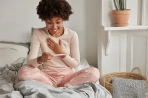 Cheerful African American future mother cant believe in positive result on pregnancy test, awakes early in morning, wears casual pyjamas, poses against bedroom interior